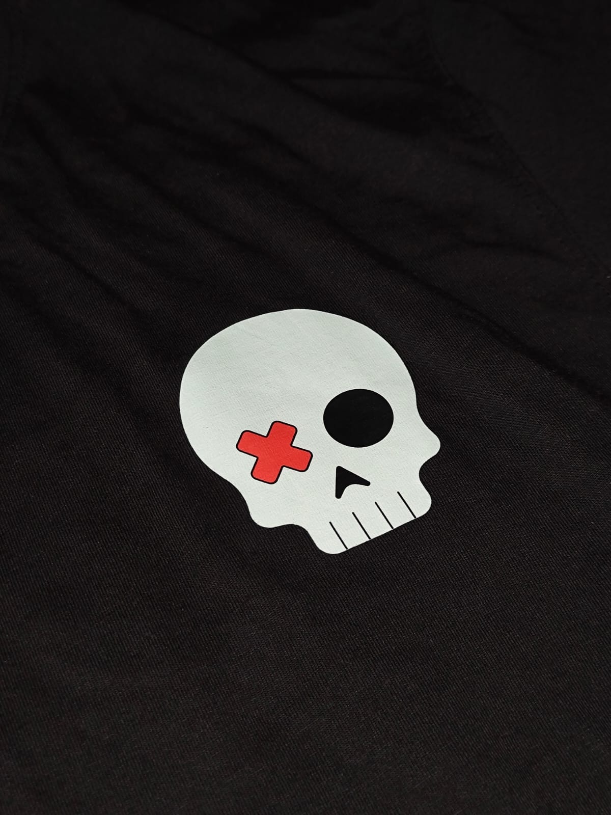 A close-up of a black fabric garment featuring a white skull graphic with a red cross on one eye and a black eye socket.